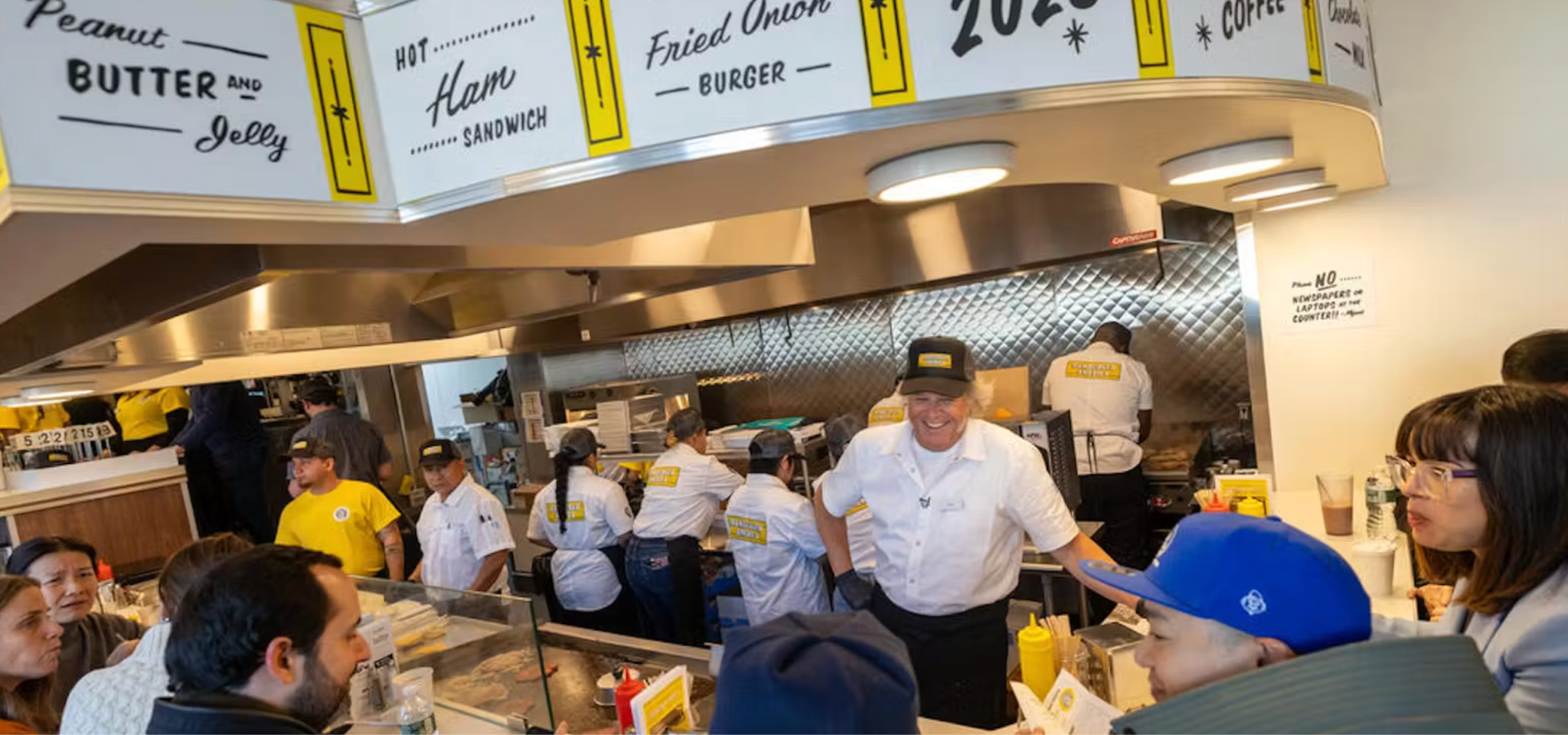 Retro signage hangs above the open kitchen. Visitors sit around the kitchen, waiting for their burger orders.