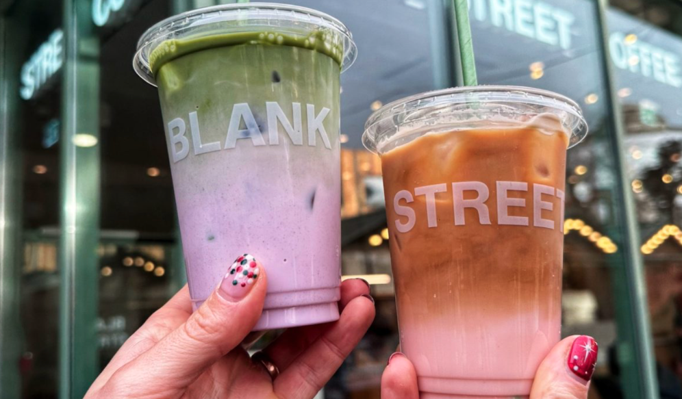 Consumers hold two lattes, one green and purple and one brown and pink, outside of a Blank Street coffeehouse. 