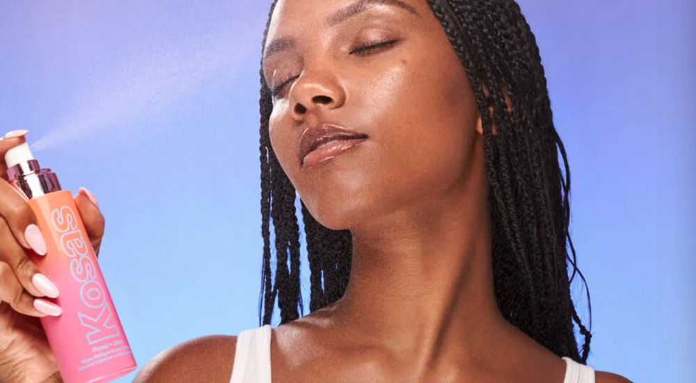 Woman sprays her face with a bottle of Kosas setting spray. 