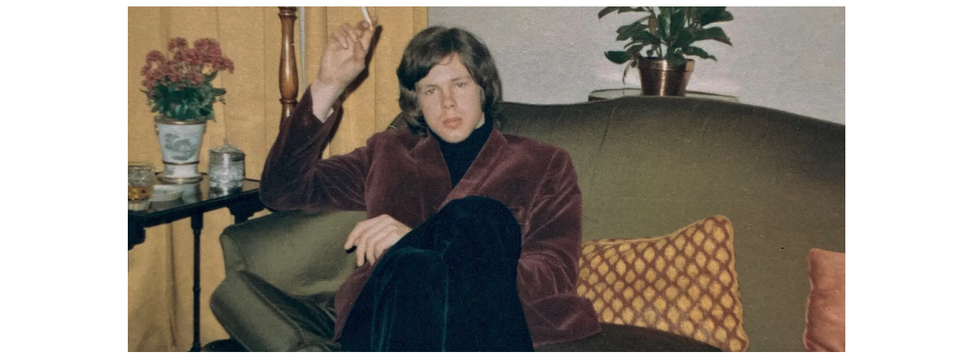 Nick Drake sits on a green sofa, he wears a brown jacket and green velvet trousers and holds a cigarette in one hand. 
