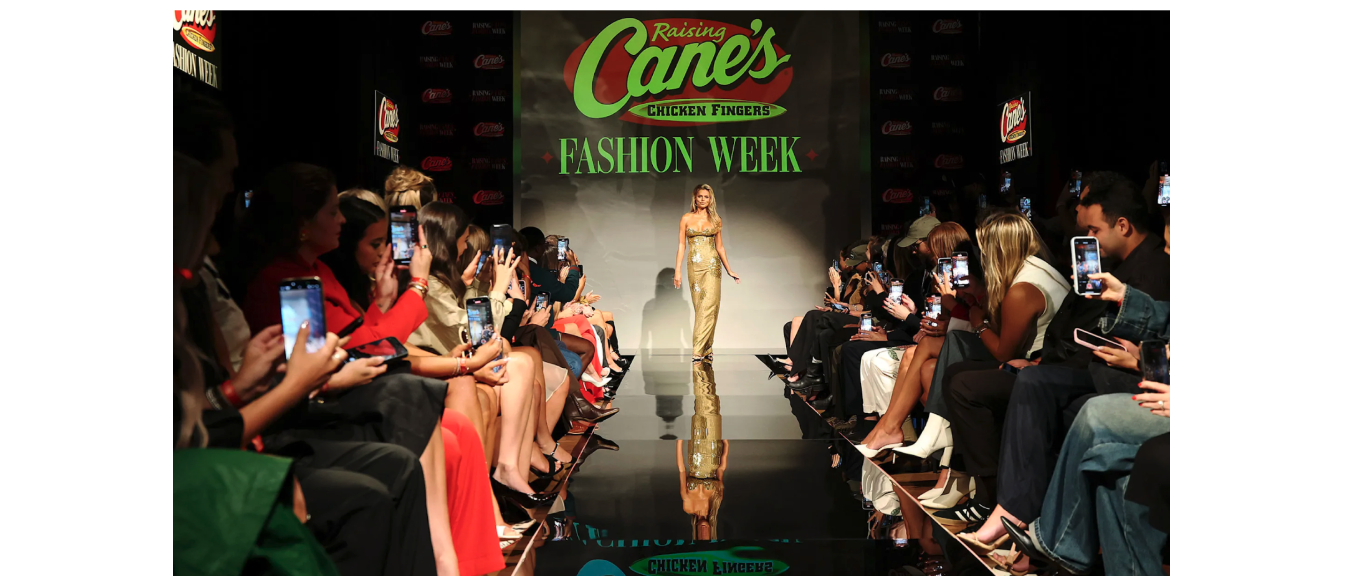 The model stands in a gold dress at the start of the runway, under a sign that reads, “Raising Cane’s Chicken Fingers Fashion Week.” 