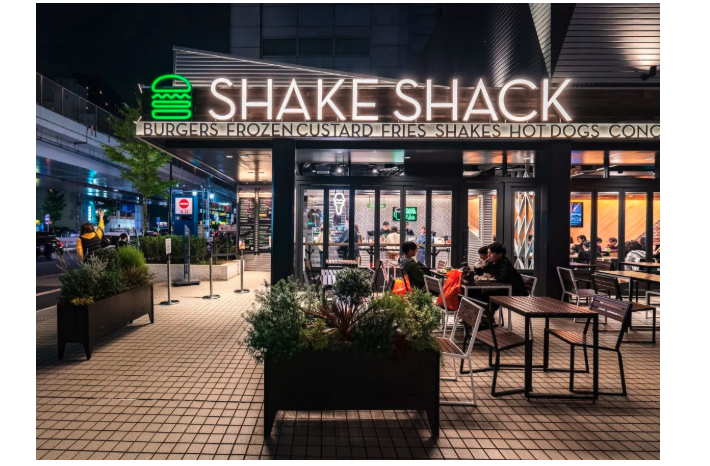 Photo of the outside of a Shake Shack location, with outdoor seating, a neon sign with a burger outlined in green, and the words “Shake Shack.”