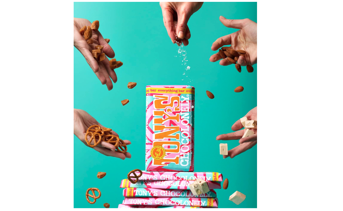 Tony’s Everything Bar sits stacked, surrounded by hands throwing salt, almonds, caramel, pretzels and nougat pieces. 