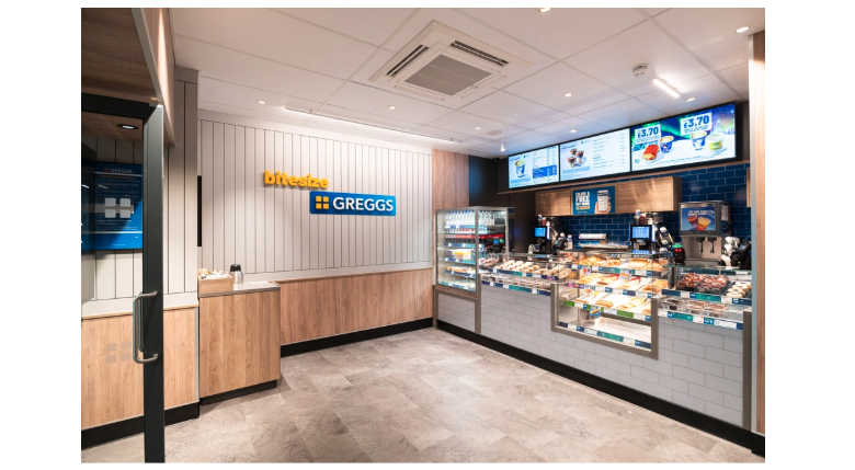 Photo of the interior of a small-format Greggs, with a counter and display case along one wall, and the brand’s logo on another.